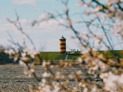 Pilsumer Leuchtturm: The photo captures a vibrant red and yellow lighthouse positioned on a green hill, framed by blooming tree branches in the foreground. The scene is set against a clear blue sky, with several people walking along the path towards the lighthouse.