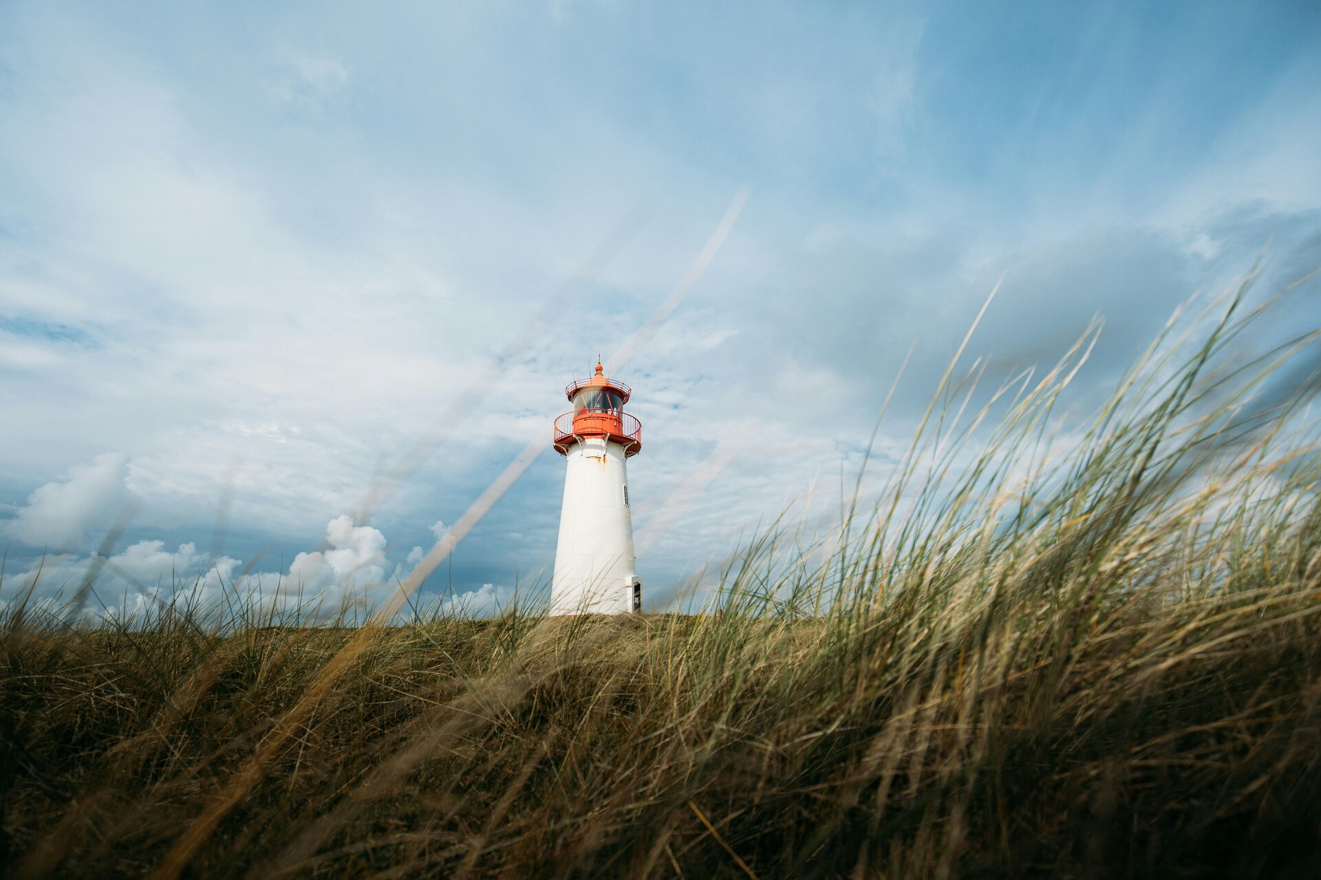 Fotospot Leuchtturm List-West Sylt: White lighthouse on grassy hill under dramatic cloudy sky.